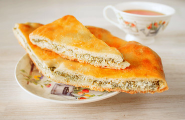 Oriental pastry fresh hot traditional Ossetian pie with homemade cheese, spinach and herbs cut into pieces in saucer with cup of tea. Close-up, selective focus.