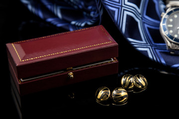 Golden cufflinks lying on a black reflective surface with a leather bound presentation box and a blue tie and stylish watch in background