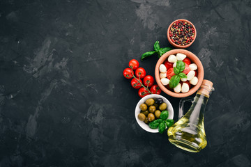 Cooking a caprese salad. Mozzarella cheese, cherry tomatoes, olives, basil leaves, oil, pepper. On a black stone table. Free space for text.