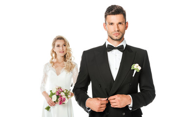 handsome young groom buttoning jacket and looking at camera with bride standing blurred on background isolated on white