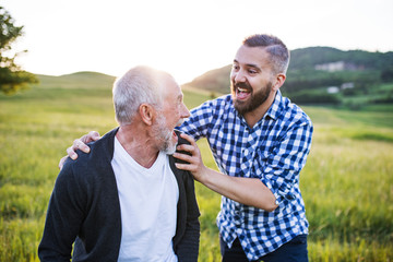 An adult hipster son with senior father on a walk in nature at sunset, having fun.