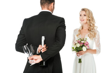 rear view of groom holding champagne bottle and glasses behind back while bride standing in front of him isolated on white