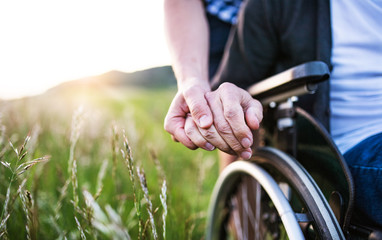 A close-up of unrecognizable son holding his father's hand on a wheelchair.