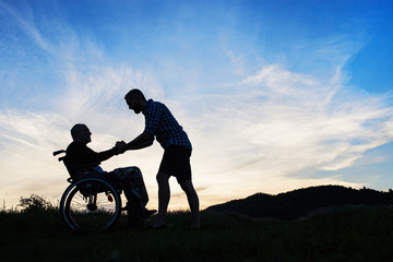 A silhouette of adult son looking at his father in wheelchair in nature at sunset.