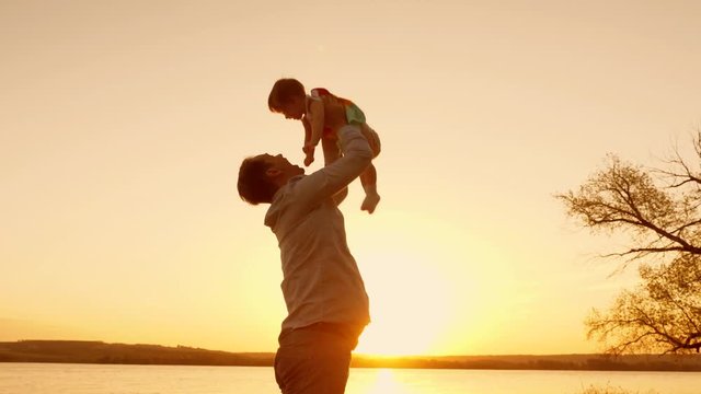Dad plays with his daughter at sunset, raises his little daughter high up to sky with his hands. Slow motion