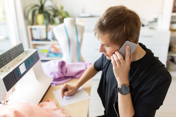 people, clothing and tailoring concept - fashion designer calling on smartphone and making notes at studio