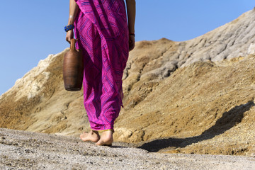 a barefooted woman in ethnical clothes carries in her hand a clay jug