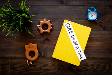 Reading at night for children. Fairy tale book near printed text once upon a time and toys on dark wooden background top view