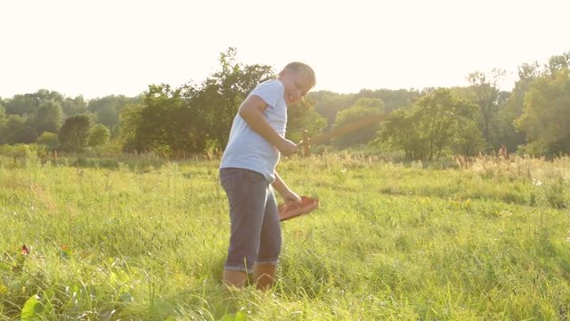 Young white healthy kid cheerfully playing with wooden toys outside in countryside meadow landscape on sunny sunset summer evening. Boy holds sword and shield. Real time full hd video footage.