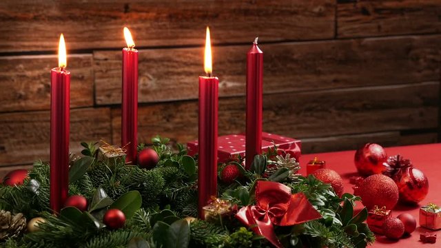 Three burning red candles on a traditional advent wreath of green fir twigs and mistletoes with festive decoration in front of a rustic wooden wall, close-up real time shot with copy space, nobody