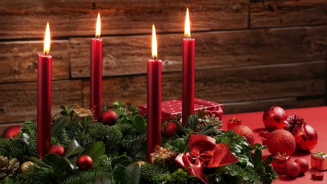 Four burning red candles on a traditional advent wreath of green fir twigs and mistletoes with festive decoration in front of a rustic wooden wall, close-up real time shot with copy space, nobody