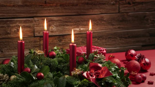Four burning red candles on a traditional advent wreath of green fir twigs and mistletoes with festive decoration in front of a rustic wooden wall, close-up time lapse shot with copy space; approx. 27