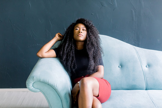 Young African American girl in black tshirt and red trousers sitting on a blue sofa on a dark background