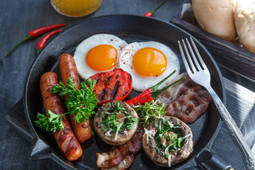 Close view of English Breakfast in a pan with fried eggs, sausages, bacon, mushrooms, jam and orange juice on dark stone background
