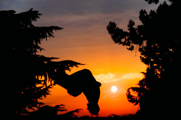 Fototapeta na wymiar Silhouette of young parkour man while jumping and flipping upside down in park on a sunrise. 