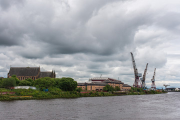 Glasgow, Scotland, UK - June 17, 2012: Along River Clyde and harbor, cranes and warehouse roofs along with Govan Old Parish church at left. Dark gray water and heavy rainy cloudscape.