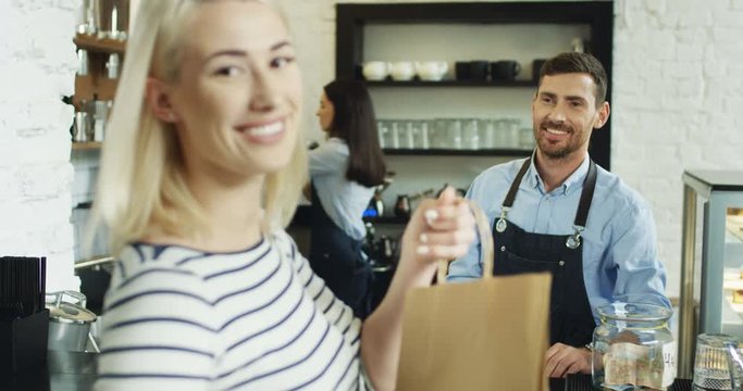 Beautiful blonde woman buying something in the cafe and good looking smiled waiter giving her a cartoon bag at the bar.