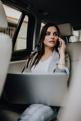 Young woman with laptop sitting in car and talking on the phone