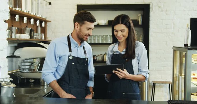 Attractive Caucasian waitress and waiter smiling and watching something on the tablet computer while standing at the bar in the cafe.