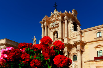 Flores rojas floreciendo en primer plano. Detrás fachada principal de una iglesia. Fotografía tomada en Valetta, Malta. Cielo azul como fondo. 