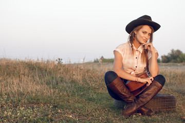 Girl cowgirl in jeans boots shirt and with a bag in a box with a wheel by a tree