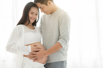 Handsome Asian man and his beautiful pregnant wife are hugging and smiling while standing near the window at home.