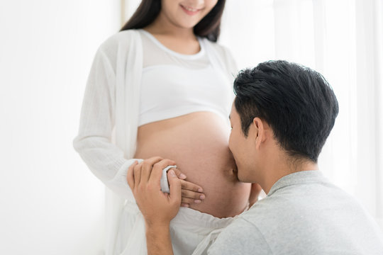 Asian Husband Kissing Pregnant Belly Of His Wife With His Eyes Closed. Asian Married Couple And Family Concept.