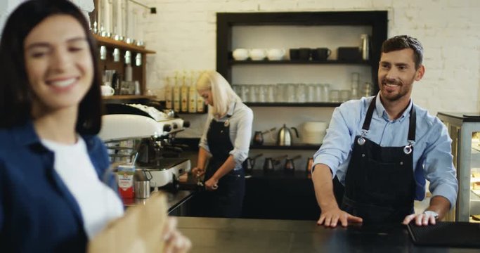 Brunette woman buying something in the cafe and handsome smiled waiter giving her a cartoon bag at the bar, then she going away.