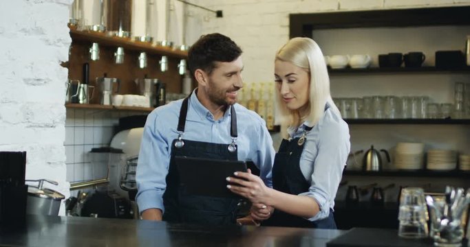 Cheerful good looking waiter and waitress standing at the bar, talking and watching some video on the tablet computer.