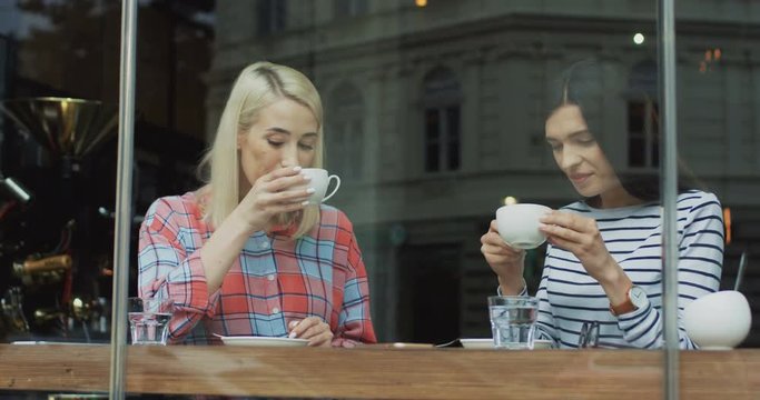 Two good looking young woman sitting in the cafe behind the window and drinking coffee.