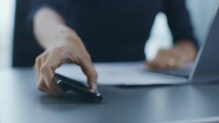 Out of Focus Shot of the Businesswoman Working at Her Office Desk Reaching Out for Her Smartphone and Starts Using It. Woman Picks up Mobile Phone from Her Desk. Focus on a Phone. - Powered by Adobe