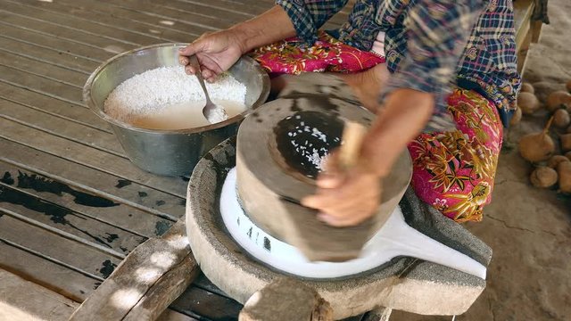 woman sat cross-legged using hand-turned millstone to grind wet rice for making soaked rice flour
