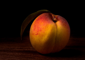 Peach still life one fruit seen from the side against black background highlighting the golden colors and texture and with one leaf