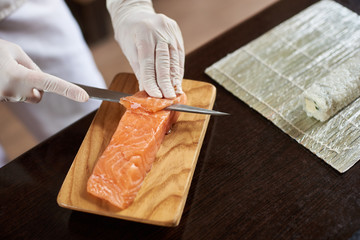 Close-up view of process of preparing delicious rolling sushi in restaurant. Female hands in disposable gloves slicing salmon on wooden board.