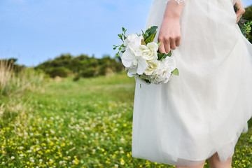 Asian woman wearing white laced bridal dress  holding white bouquet at her side at the yellow blossom field located on the cliffs of the beautiful Jeju Island, Korea.           