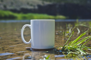 A solo blank white coffee mug on the shores of the green river in the bright morning summer sun. 