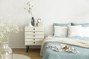 A bright bedroom interior with sage green and white bedding, pillows on bed and a drawer...