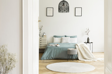 A view through an open door into a sunny bedroom interior with sage color linen and cushions on a bed, blanket, drawer cabinet, soft stool and a round rug. Real photo