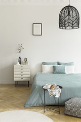 Breakfast on a metal table in front of a bed with sage green bedding in a natural bedroom interior. A beige drawer cabinet by the bed. Black lamp hanging from a ceiling. Real photo