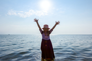 Child girl in summer dress with straw hat is standing barefoot in shallow sea water with raised hands