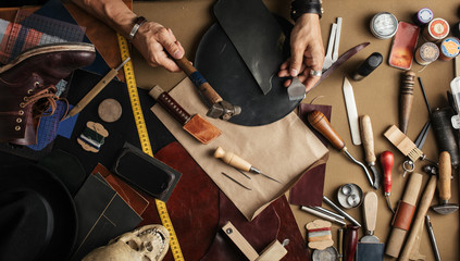 High angle view of craftsman at work over background with set of cobbler tools and colored leather in rolls. Genuine leather handbag master at work in local workshop. Handmade concept. - obrazy, fototapety, plakaty