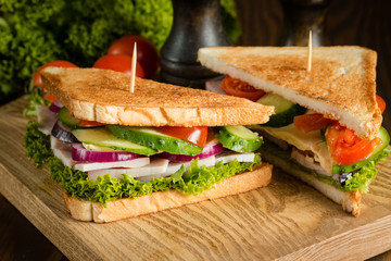 Close-up of two sandwiches with bacon, salami, prosciutto and fresh vegetables on rustic wooden cutting board. Club sandwich concept.
