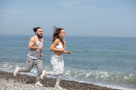 Happy caucasian athletic couple running together beside the ocean water at the pubble beach in the sunny summer day.