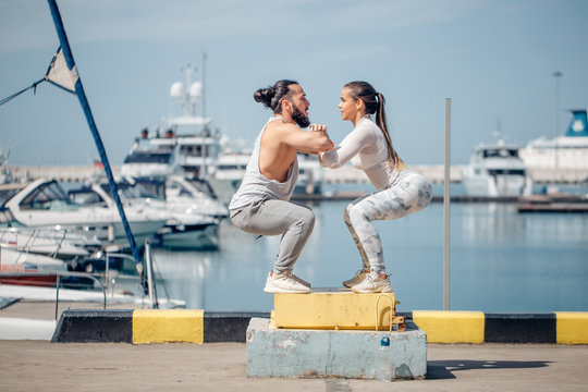 Fit young woman and athletic beared man box jumping in a crossfit style at the ocean pier with marinas. Female and male athlete is performing box jumps outdoor.