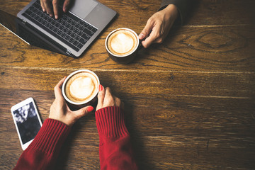 Female hand holding cup of hot latte coffee and mobile phone with friend working on laptop at coffeeshop, co working space concept