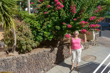 Aged woman is standing under bougainvillea bush.