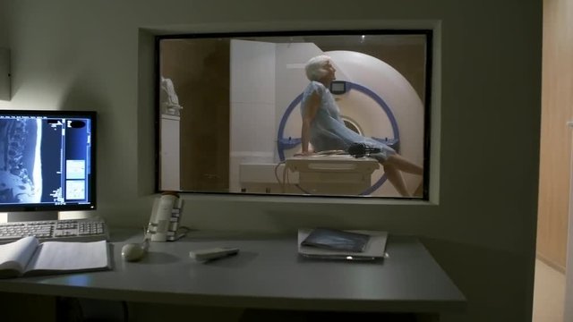 Panning shot of MRI exam room with desk and computer on it; senior woman in hospital gown sitting on medical couch, swinging legs and waiting for procedure