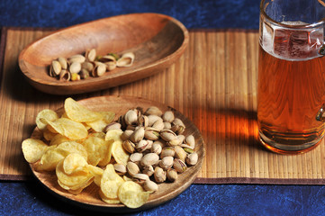 A mug of light beer on a napkin made of bamboo. Nearby snacks are pistachios in a bowl and potato chips on a plate. Dark background. Close-up. View from above.