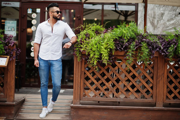 Stylish tall arabian man model in white shirt, jeans and sunglasses posed at street of city. Beard attractive arab guy.