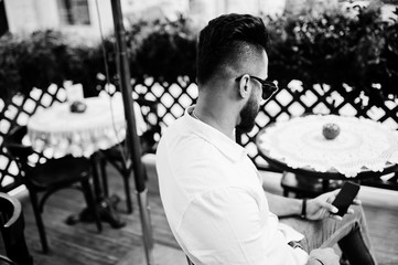 Stylish tall arabian man model in white shirt, jeans and sunglasses posed at street of city. Beard attractive arab guy sitting on outdoor cafe and looking at mobile phone.